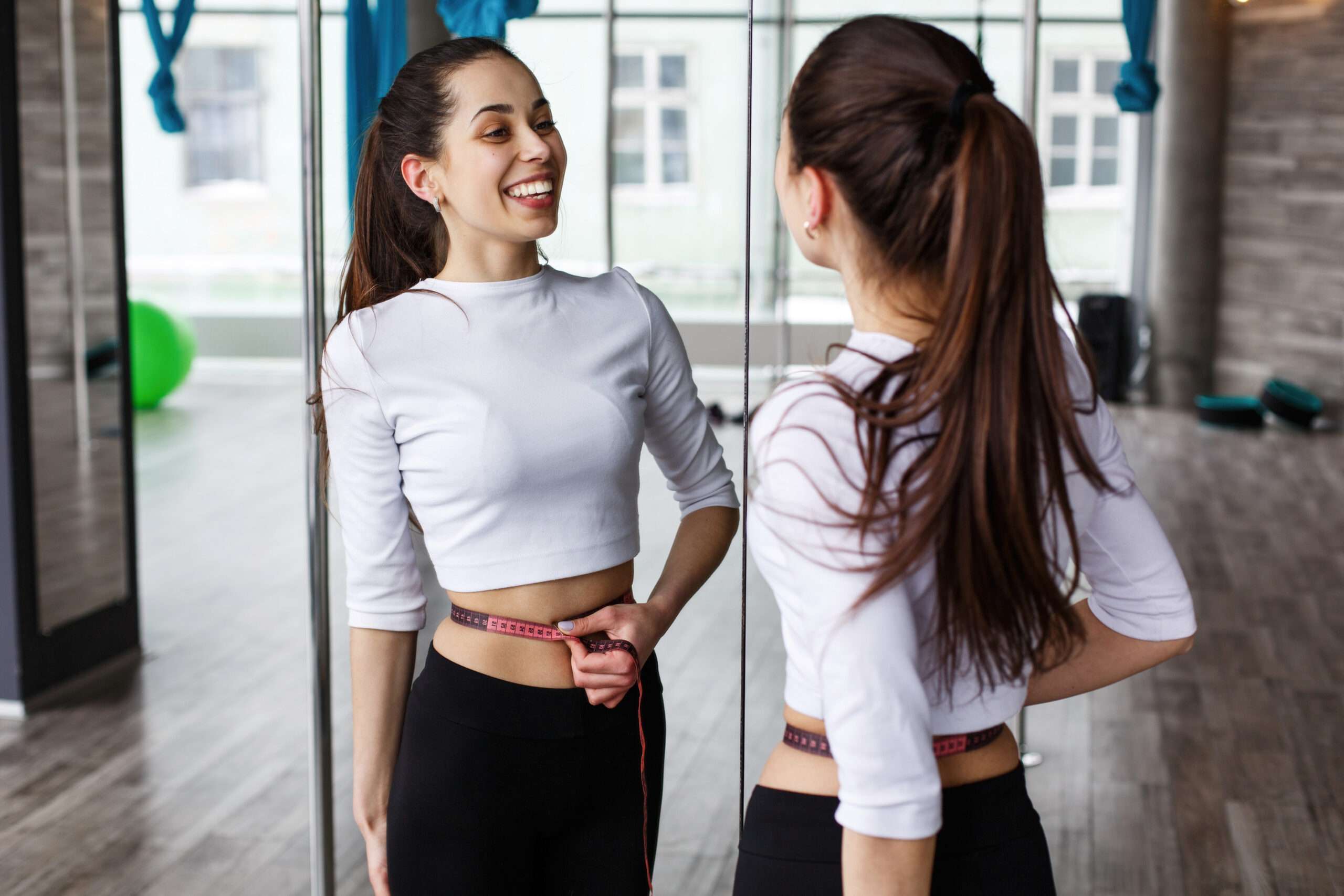 Happy woman looks at herself in the mirror holding a tape-measur Jeera Paani Peene Ke Fayde